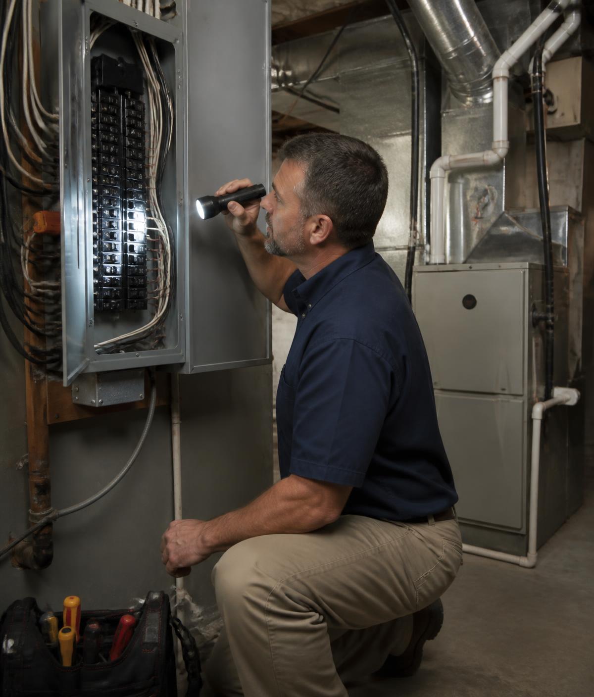 Home inspector using a flashlight to examine an electrical panel and furnace in a residential basement